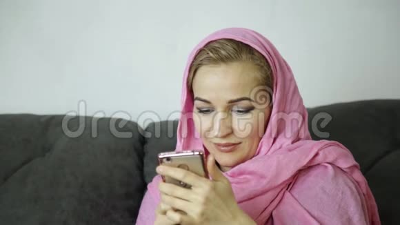 穿着粉红色头巾的年轻穆斯林妇女坐在咖啡馆的沙发上用手机发短信视频的预览图
