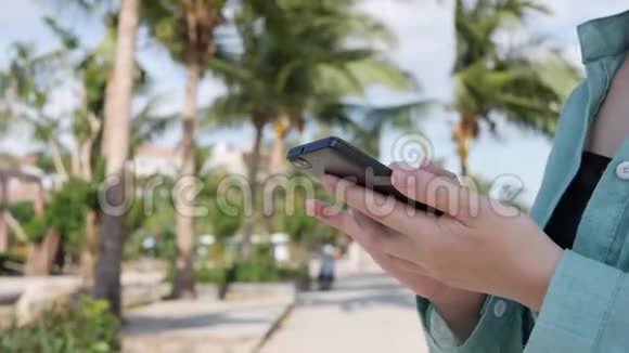 美丽的苗条女人一头金色长发穿着绿色衬衫站在棕榈树附近在后台使用智能手机视频的预览图