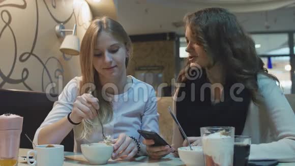 两个漂亮的年轻女孩坐在咖啡馆里吃着看着手机屏幕视频的预览图