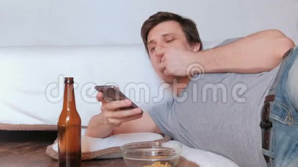 英俊的年轻人单身汉吃薯片喝啤酒浏览他的手机视频的预览图