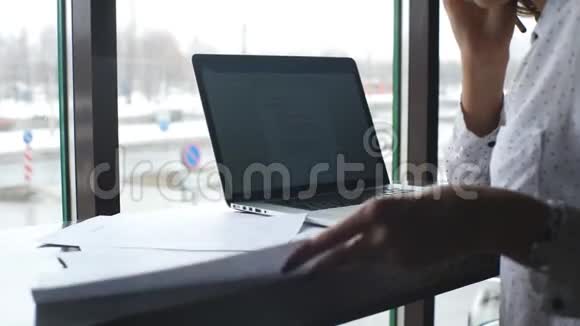 这位年轻的女商人在一家带着笔记本电脑的咖啡馆工作在文件上签名并在电话上交谈视频的预览图