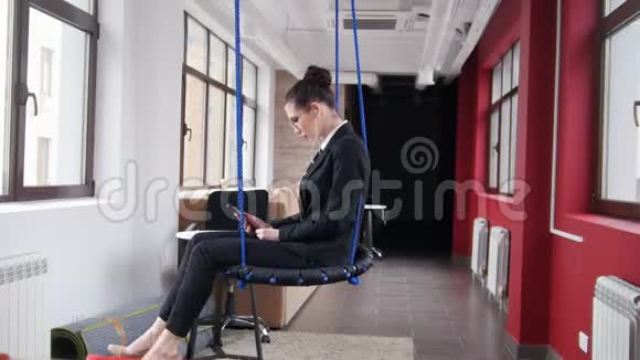 商业概念一个戴眼镜的女人坐在办公室的秋千上听音乐视频的预览图