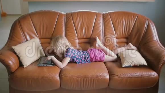 一个可爱的金发小女孩穿着所有粉红色的侧视图躺在沙发上在平板电脑上玩游戏慢动作视频的预览图
