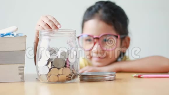亚洲小女孩把硬币放在一个透明的玻璃罐子里的桌子上比喻省钱的概念声音选择关注罐子视频的预览图