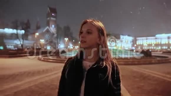 一个年轻的女孩在一个冬天的晚上在城镇广场房子的灯光下雪了女孩在等待视频的预览图