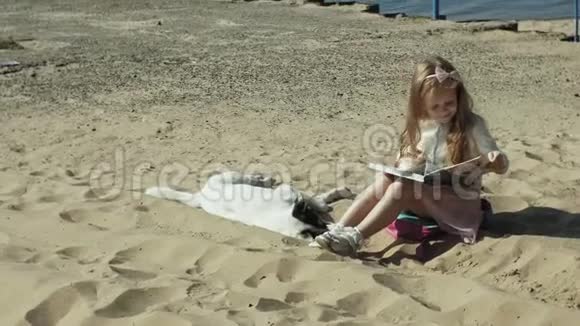 一个可爱的女孩坐在沙滩上读一本书抚摸一只狗视频的预览图