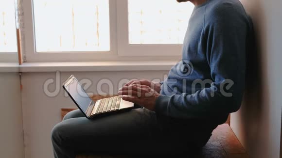 在笔记本电脑上工作不可辨认的年轻人打开笔记本电脑等待它启动坐在阳台附近的窗户视频的预览图