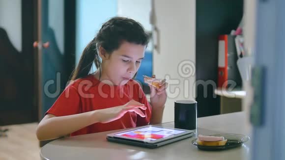 小女孩在玩数字平板电脑吃三明治坐在桌旁玩数字平板电脑的小女孩视频的预览图