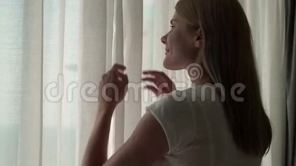年轻女人揭开窗帘的剪影望着窗外在外面欣赏海景慢动作100fps视频的预览图