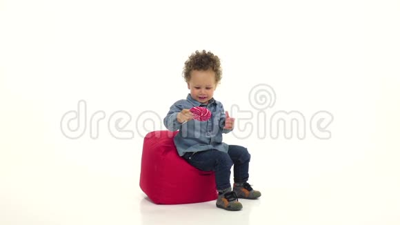 孩子是非裔美国人坐在枕头上舔糖果白色背景慢动作视频的预览图
