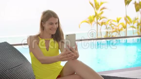 穿着黄色衣服的女人坐在靠近游泳池的日光浴床上用手机自拍视频的预览图