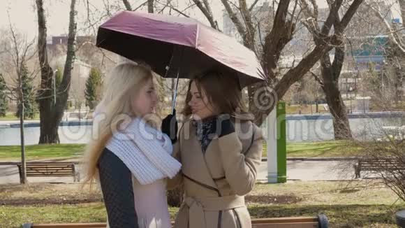 两个美丽的女朋友金发和黑发站在伞下说话晴天风吹视频的预览图