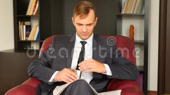 这位商人坐在椅子上看报纸从瓶子里喝酒精他不快乐很有表现力视频的预览图