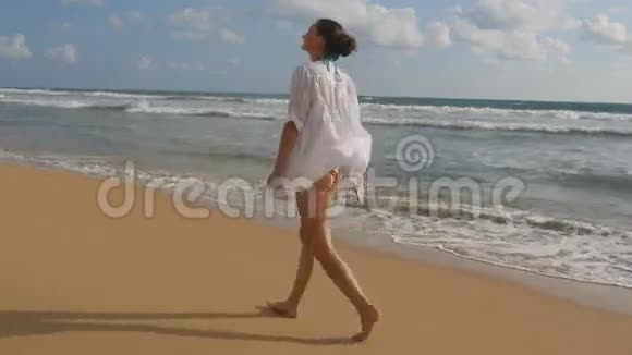 美丽的女人穿着泳衣和衬衫光着脚在海边散步举起双手小女孩在大海上视频的预览图