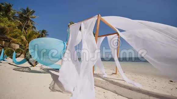 婚礼拱门在靠近海洋的海滩上装饰着鲜花她的白色和蓝色的画布在风中飘扬装饰视频的预览图