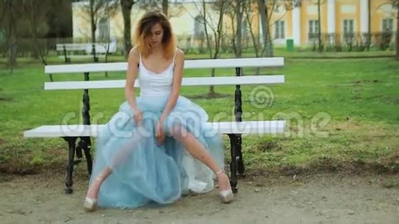 漂亮的瘦女孩穿着银色和蓝色连衣裙穿着高跟鞋坐在Parkway的长凳上膝盖磕磕碰碰视频的预览图