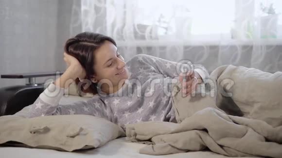 可爱的微笑的黑发女孩短发穿着灰色睡衣躺在卧室的沙发上用她的头发视频的预览图