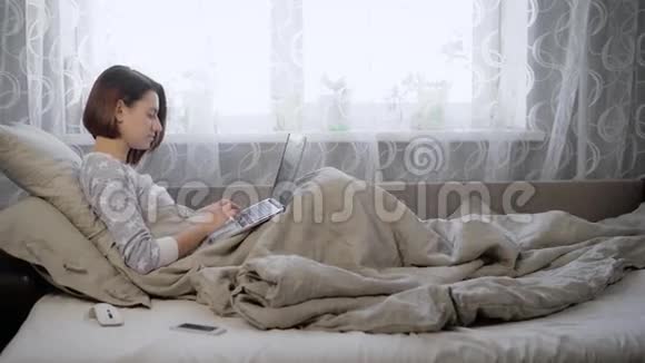 棕色头发的年轻女孩穿着灰色毛衣坐在床上毯子盖在枕头上在她身上视频的预览图