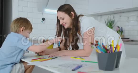 年轻漂亮的妈妈和儿子坐在厨房的桌子旁用彩色铅笔画画视频的预览图