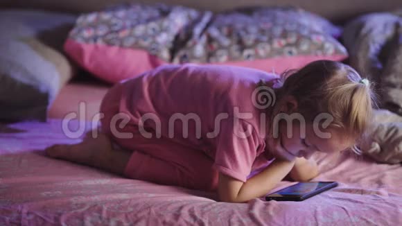 穿着粉红色睡衣手里拿着手机的小女孩躺在床上看动画片幼儿视频的预览图