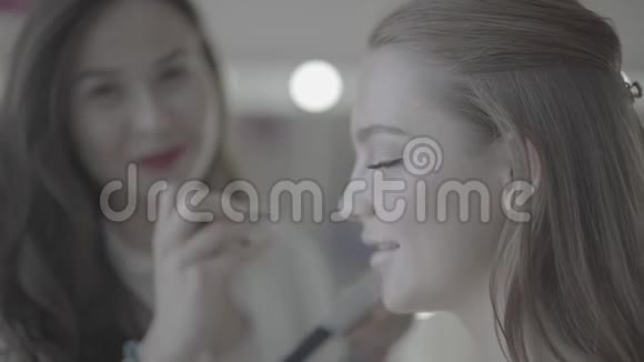 年轻漂亮的黑发女性化妆师在美容院精心制作漂亮的金发模特脸视频的预览图