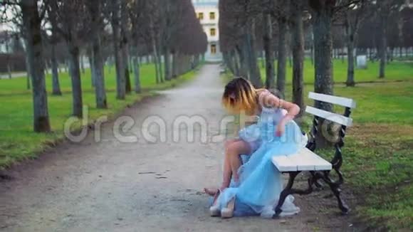 穿着银色和蓝色连衣裙的漂亮女孩坐在帕克威的长凳上穿上高跟鞋准备摆好姿势视频的预览图