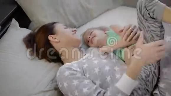 穿着时尚睡衣的快乐妈妈和女儿一起躺在床上可爱的家人在玩视频的预览图
