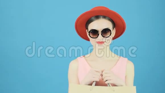 戴着红色帽子和凉爽太阳镜的可爱黑发女郎手里拿着购物袋季节性销售理念蓝色隔离视频的预览图
