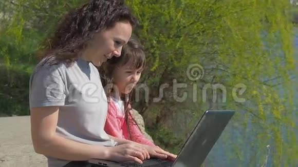 有婴儿和笔记本电脑的女人河岸上的幸福家庭妈妈在用笔记本电脑教孩子春天阳光明媚的一天视频的预览图
