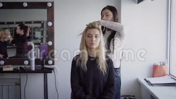 豪华美容院的年轻女美发师正站着用熨斗和年轻的金发模特一起工作美丽的视频的预览图