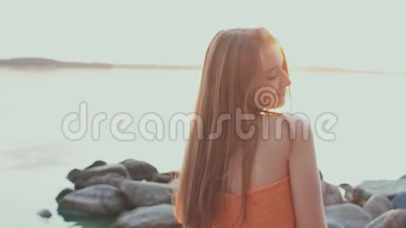一个穿着橙色连衣裙的年轻女孩在海滩上日落时摆姿势视频的预览图