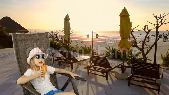 年轻的白种人女孩坐在扶手椅上用吸管喝橙汁鸡尾酒海上海岸地平线视频的预览图