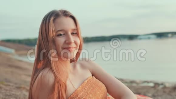 一个穿着橙色连衣裙的年轻女孩在海滩上日落时摆姿势视频的预览图