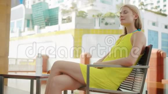 穿着黄色衣服坐在城市游泳池附近的椅子上的漂亮梦幻的年轻女子视频的预览图