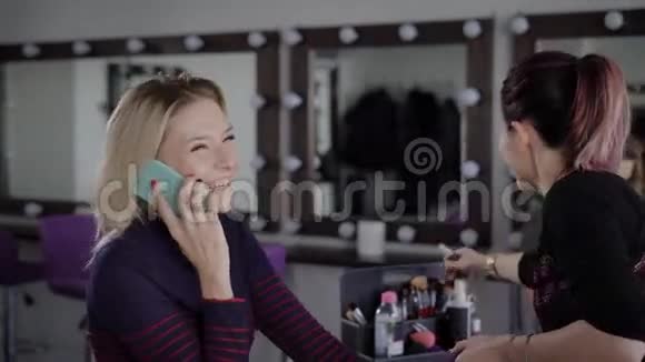 化妆师把头发缩在尾巴上为一个在时髦电话上说话的女孩做海绵眼遮瑕膏视频的预览图