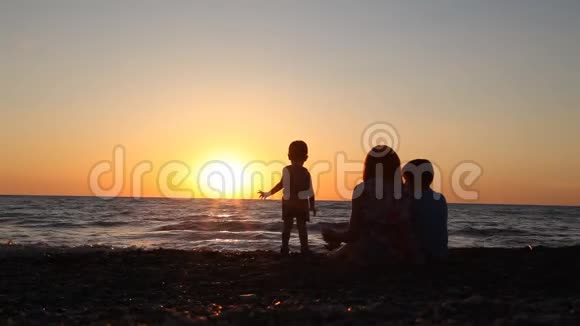 母亲的剪影两个男孩坐在海滩上儿子在日落时出海视频的预览图