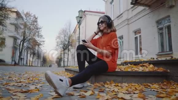 可爱的少女听耳机音乐都市风格时髦的时髦少女坐在城市街道的人行道上视频的预览图