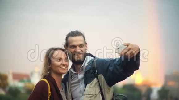 年轻的幸福夫妇在背景上拍摄彩虹的自拍照片微笑的男人和女人使用智能手机视频的预览图