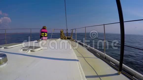 年轻的女孩坐在白色游艇的钢甲板上穿过蓝色的水阳光和蓝天视频的预览图