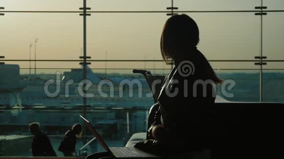 在机场候机楼用笔记本电脑工作的年轻女子在等我的航班在一个大的背景下剪影视频的预览图