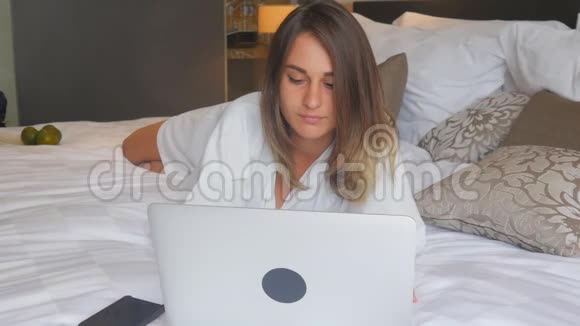 穿着浴袍躺在床上手提电脑工作的年轻女子视频的预览图