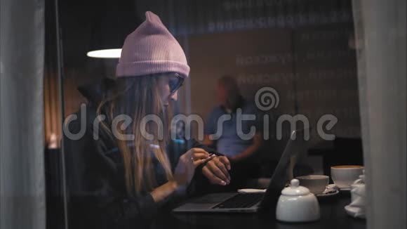 穿着时髦眼镜和帽子的嬉皮士女人坐在咖啡馆里用笔记本电脑打字从窗口观看视频的预览图