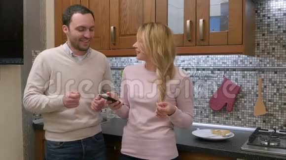 美丽的一对夫妇在家中的厨房里通过智能手机边跳舞边听音乐边笑视频的预览图
