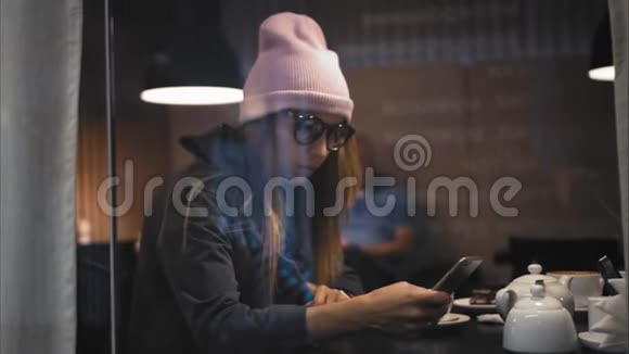 一个时髦的时髦女性戴着眼镜在咖啡馆里使用智能手机上的智能手表和应用程序从窗口观看视频的预览图
