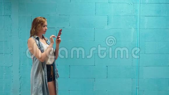 漂亮的金发女孩用红色智能手机对着蓝墙视频的预览图