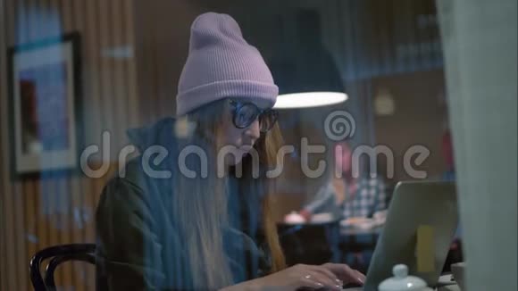 一个戴着眼镜和帽子的时髦女人坐在咖啡馆里用笔记本电脑打字从窗口观看视频的预览图