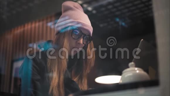 一个戴着眼镜和帽子的时髦女人坐在咖啡馆里用笔记本电脑打字从窗口观看视频的预览图