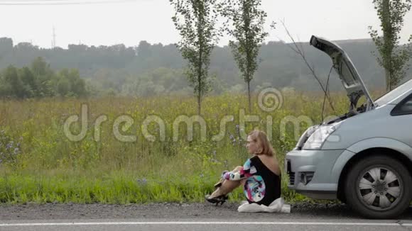 一个美丽的女孩坐在乡间小路上的破车旁打电话给手机视频的预览图