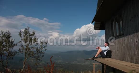 伟大的孤独英俊的年轻人坐在山顶上一座古老木屋的门廊上望着天空视频的预览图