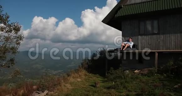 伟大的孤独英俊的年轻人坐在山顶上一座古老木屋的门廊上望着天空视频的预览图